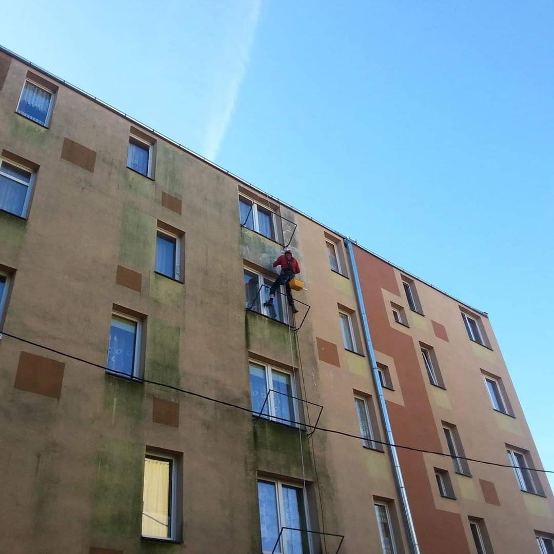 Osoba siedzi na parapecie przy oknie, w tle elewacja budynku z niebieskimi 

balustradami.